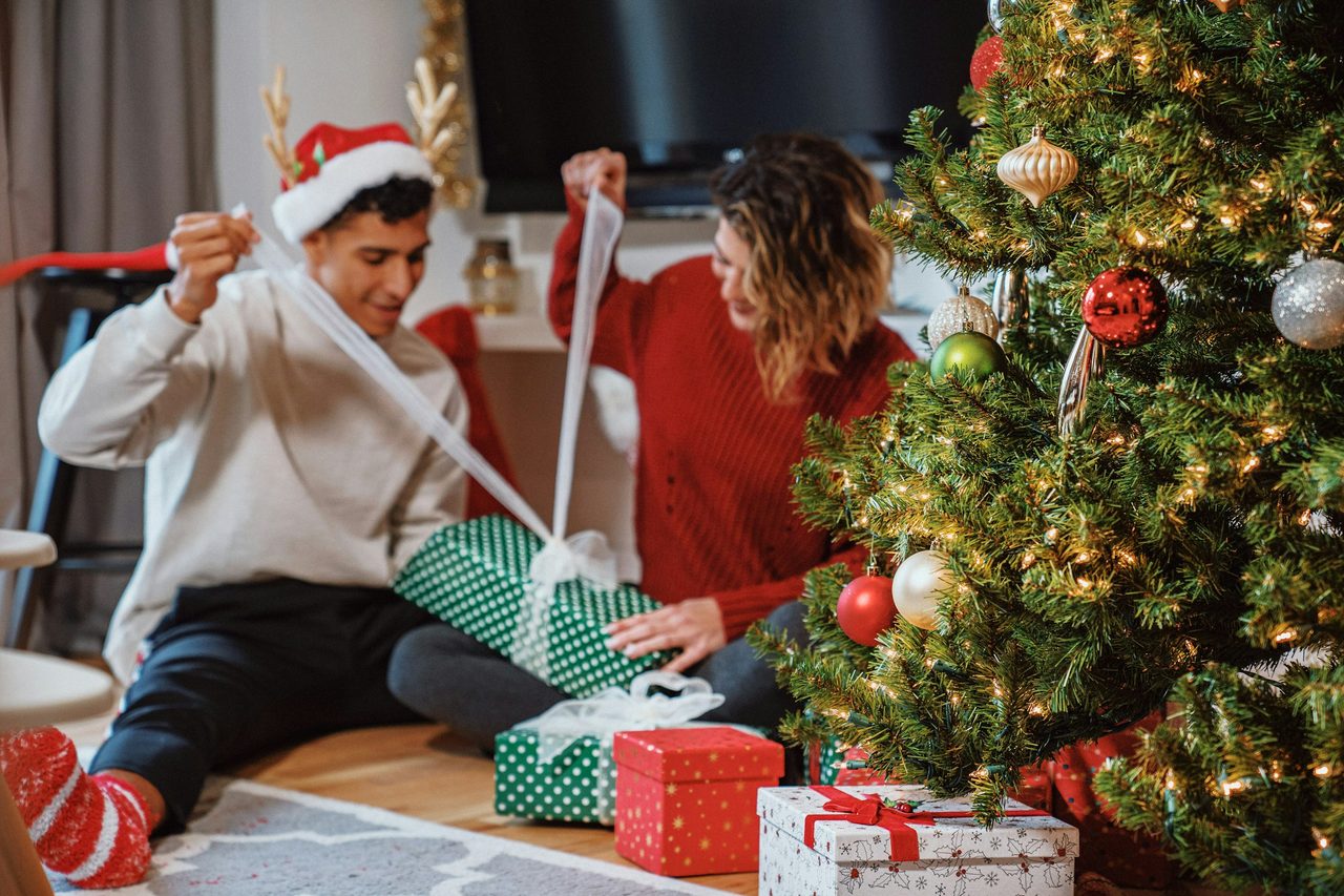 A young man and a woman pull the ribbons off wrapped presents beside a lit Christmas tree