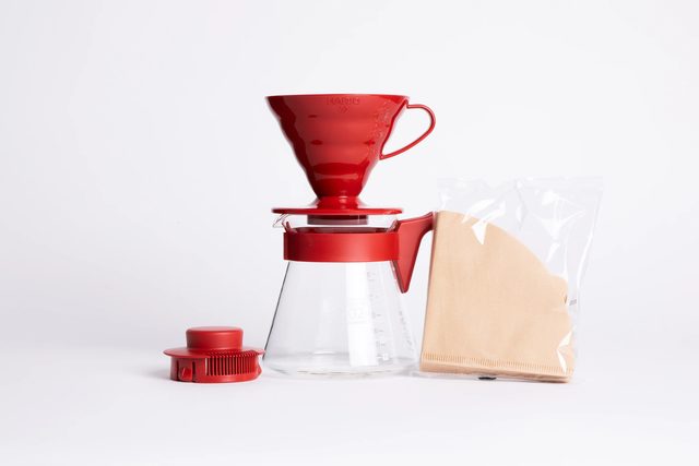 A red Hario V60 plastic dripper on a glass server with a red lid, next to a pack of paper filters