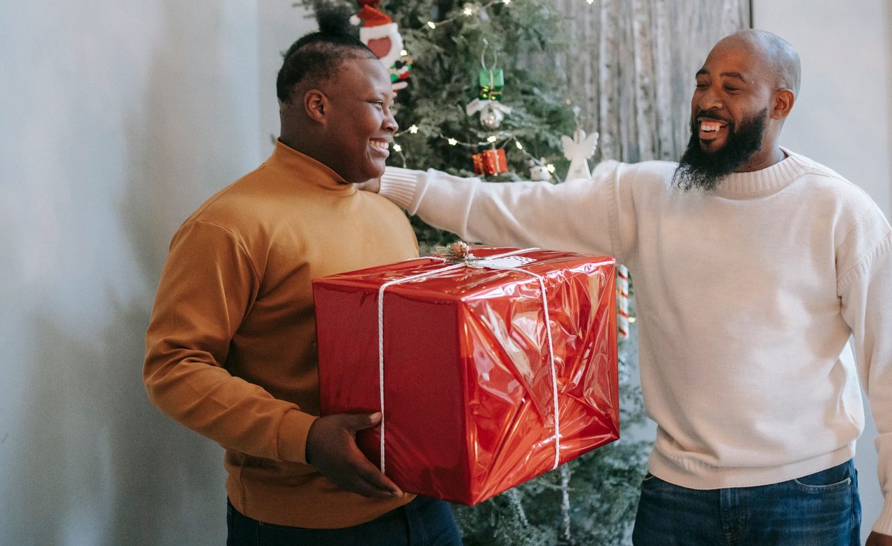 A smiling man hands his grown son a large present wrapped in red paper beside a Christmas tree