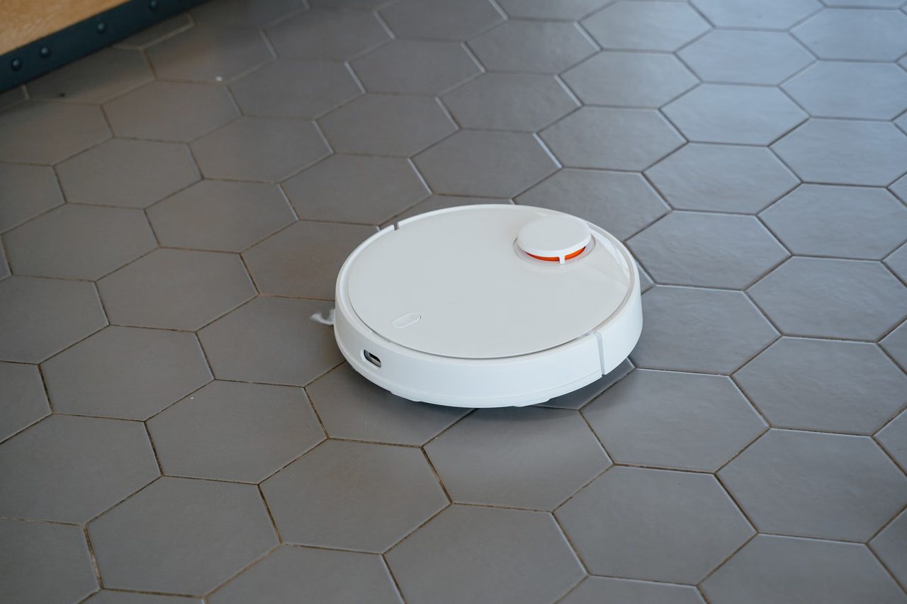 A white round robot vacuum with a LiDAR turret cleaning a gray hexagonal tile floor