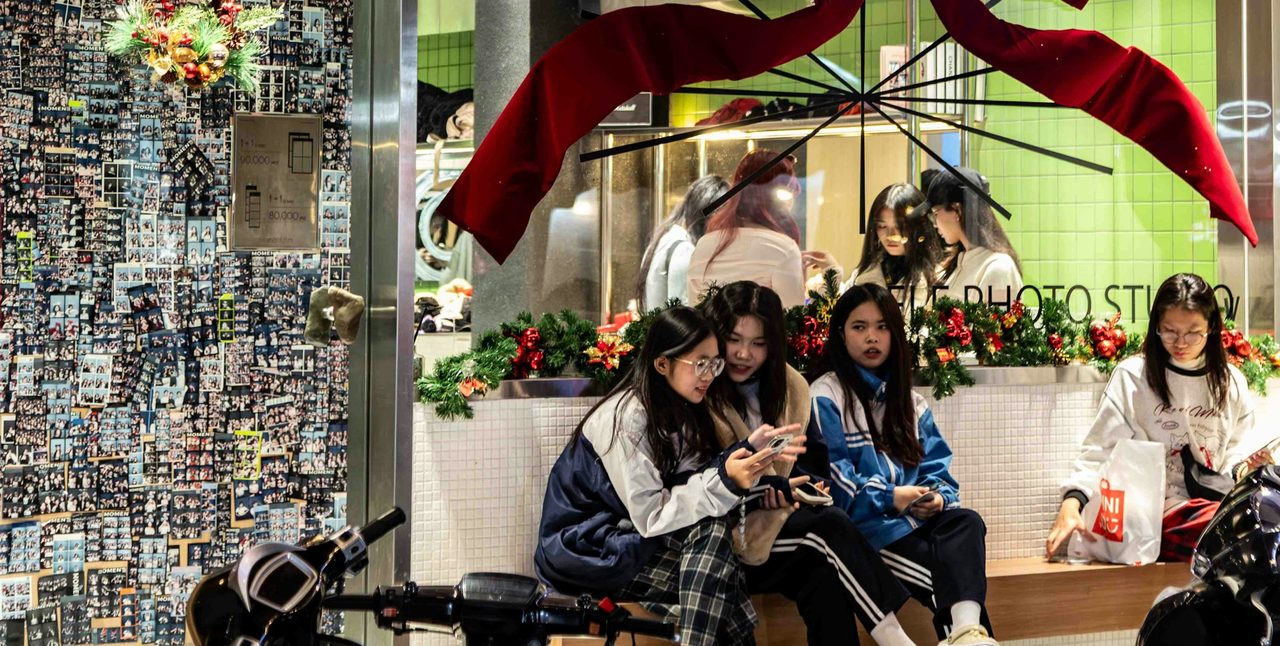 Teenage girls in school jackets sit on a bench outside a shop decorated with a giant red bow, looking at their phones