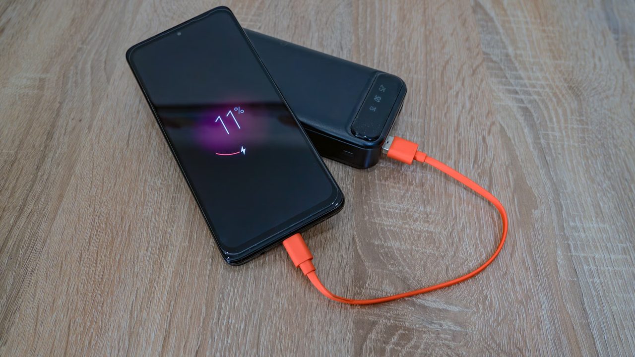 A black smartphone at 11 percent charging from a black power bank through an orange cable, on a wooden table