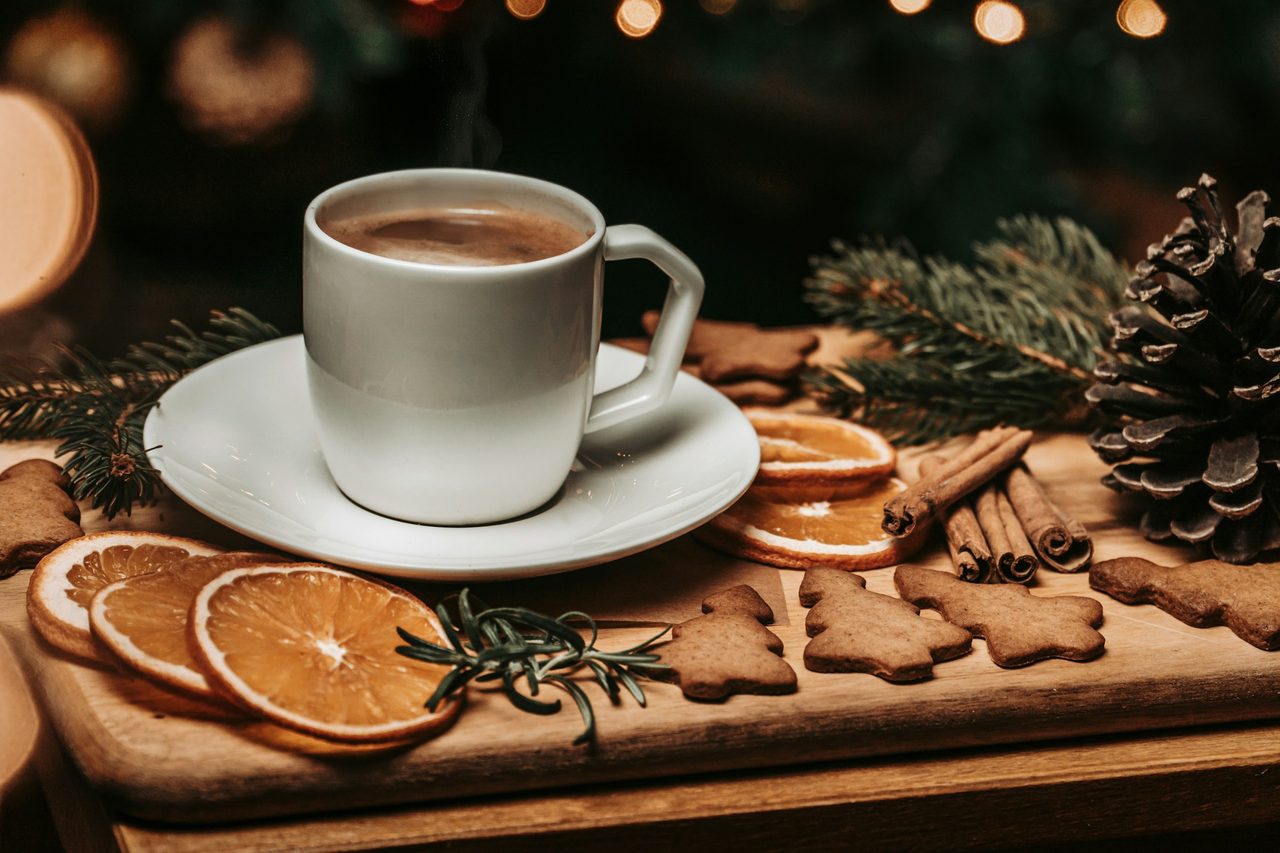 A white cup of coffee on a saucer, surrounded by gingerbread cookies, dried orange slices and pine branches