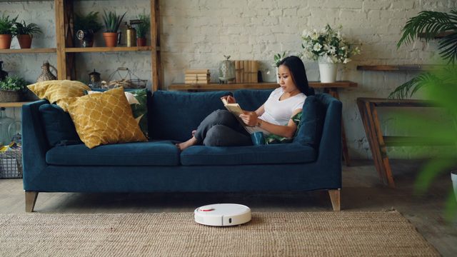 A woman reads on a blue sofa while a small white robot vacuum crosses the rug in front of her