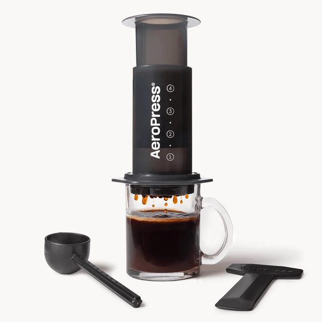 The AeroPress Original pressing coffee into a glass mug, with its scoop and stirrer beside it