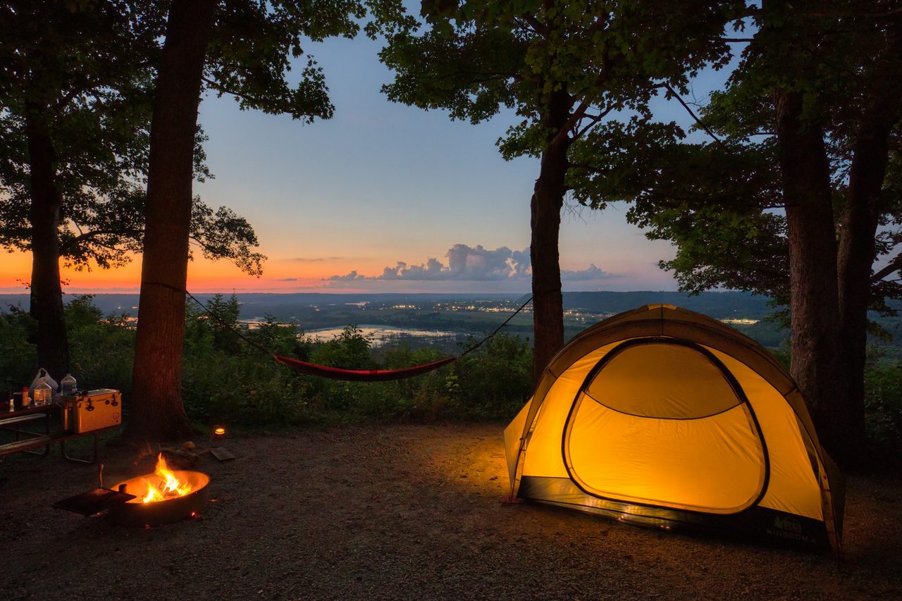 A lit dome tent beside a campfire at a wooded campsite, a lake at sunset behind it