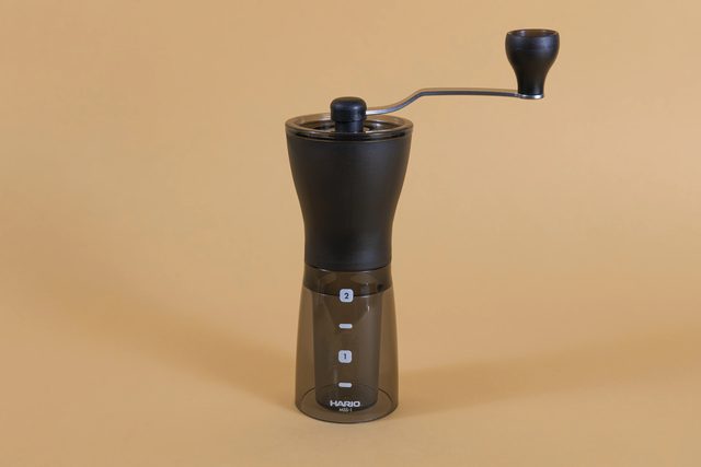 The slim black Hario Mini-Slim+ hand grinder with its crank handle on top and a clear grounds container below