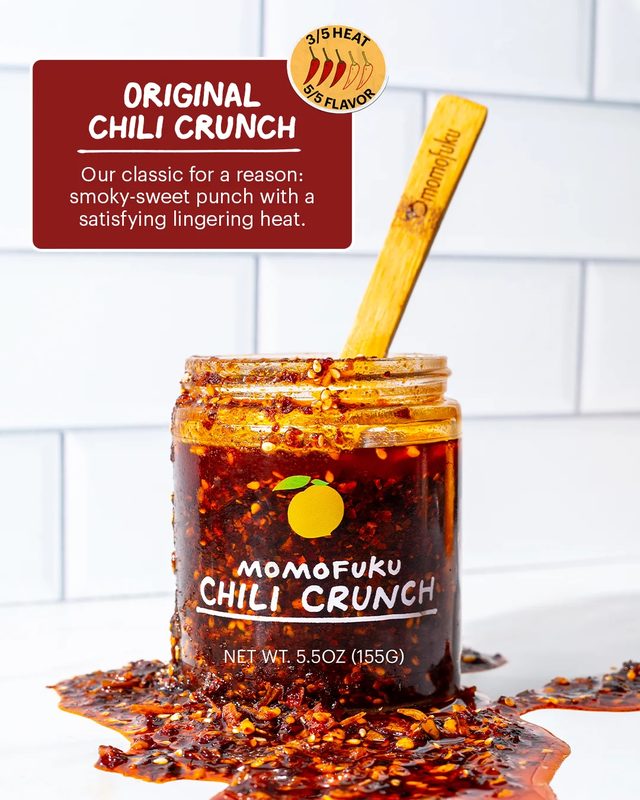 An open jar of Momofuku Chili Crunch with a wooden spoon, chili oil spilling around its base