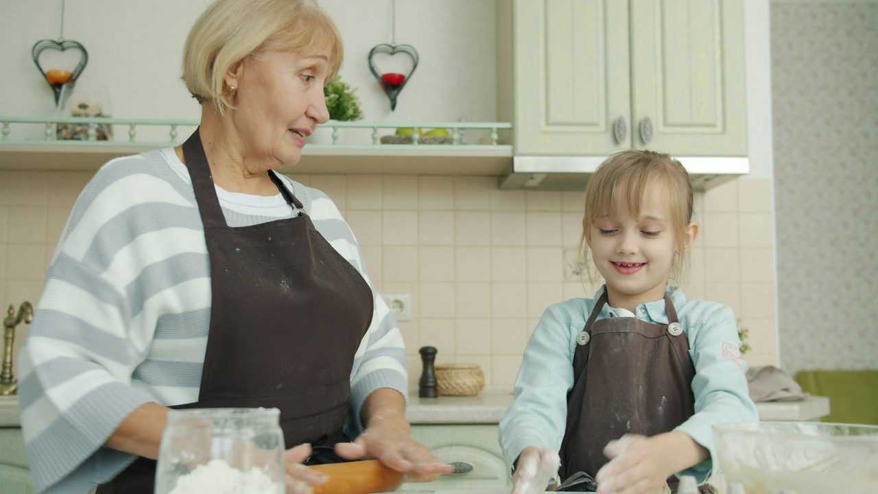 A grandmother and her granddaughter in aprons knead dough together at a kitchen counter